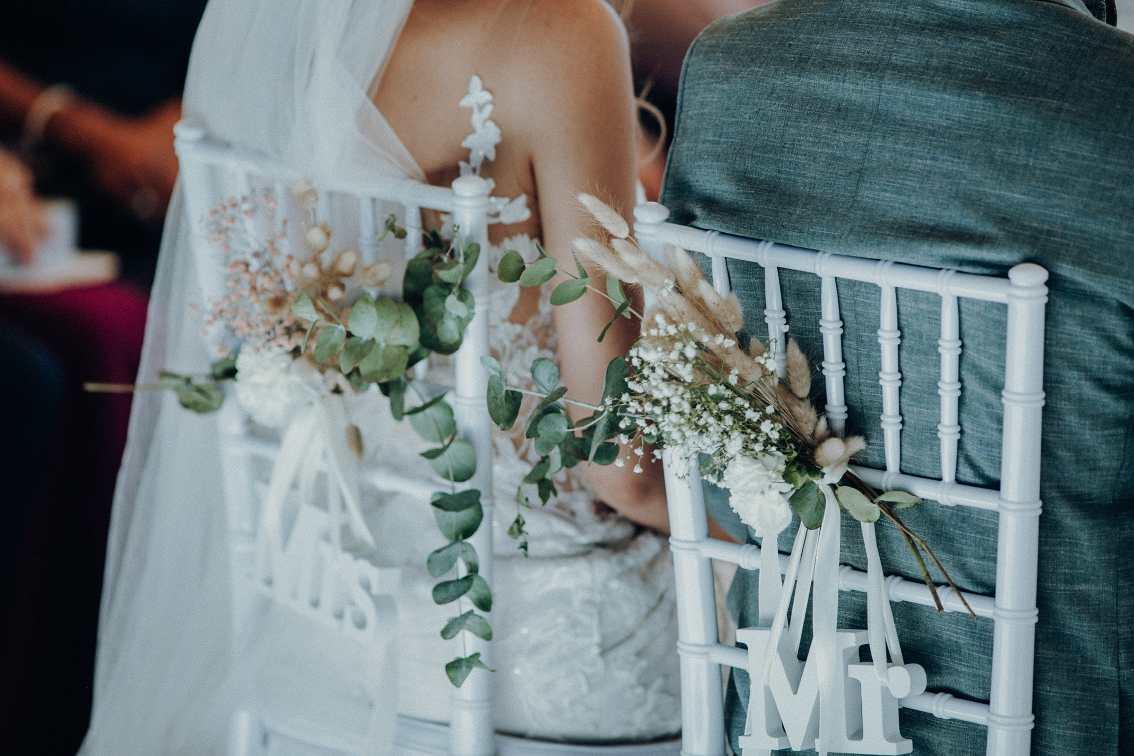 Brautpaar von hinten an der Hochzeitstafel, die Stühle mit Trockenblumen und den Schriftzügen „Mrs“ und „Mr“ geschmückt